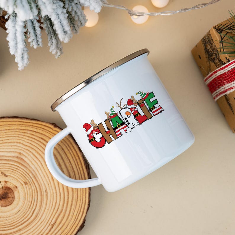 K&ouml;nnte beinhalten: Ein wei&szlig;er Emaillebecher mit silbernem Rand, der den Namen CHARLIE in gro&szlig;en, bunten Buchstaben zeigt. Jeder Buchstabe ist mit weihnachtlichen Motiven verziert, darunter Rentiere, Weihnachtsm&uuml;tzen, Streifenmuster und ein Schneemann.