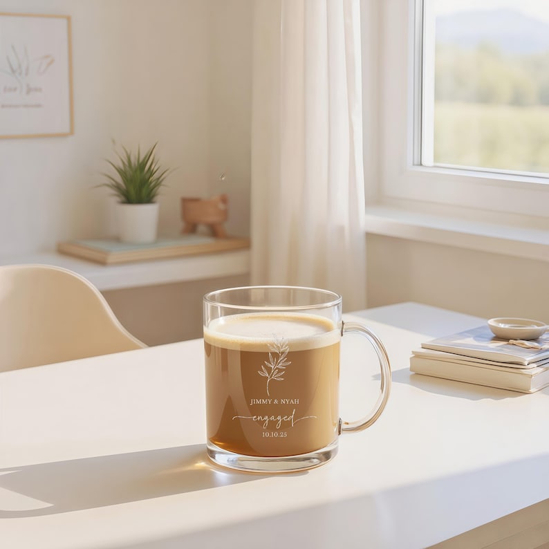 Peut inclure: Une tasse en verre transparente remplie de caf&eacute; et recouverte de mousse. La tasse a une anse et porte les mots "JIMMY & NYAH engaged 10.10.25" ainsi qu'un motif botanique. La tasse est pos&eacute;e sur une table blanche.
