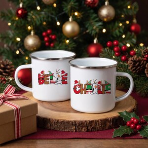 K&ouml;nnte beinhalten: Zwei wei&szlig;e Emaille-Tassen mit silbernen R&auml;ndern. Die linke Tasse zeigt den Namen BELLE in roten, gr&uuml;nen und wei&szlig;en Buchstaben im Feiertagsdesign. Die rechte Tasse zeigt den Namen CHARLIE in &auml;hnlicher festlicher Schrift mit Weihnachts- und Elfenmotiven.