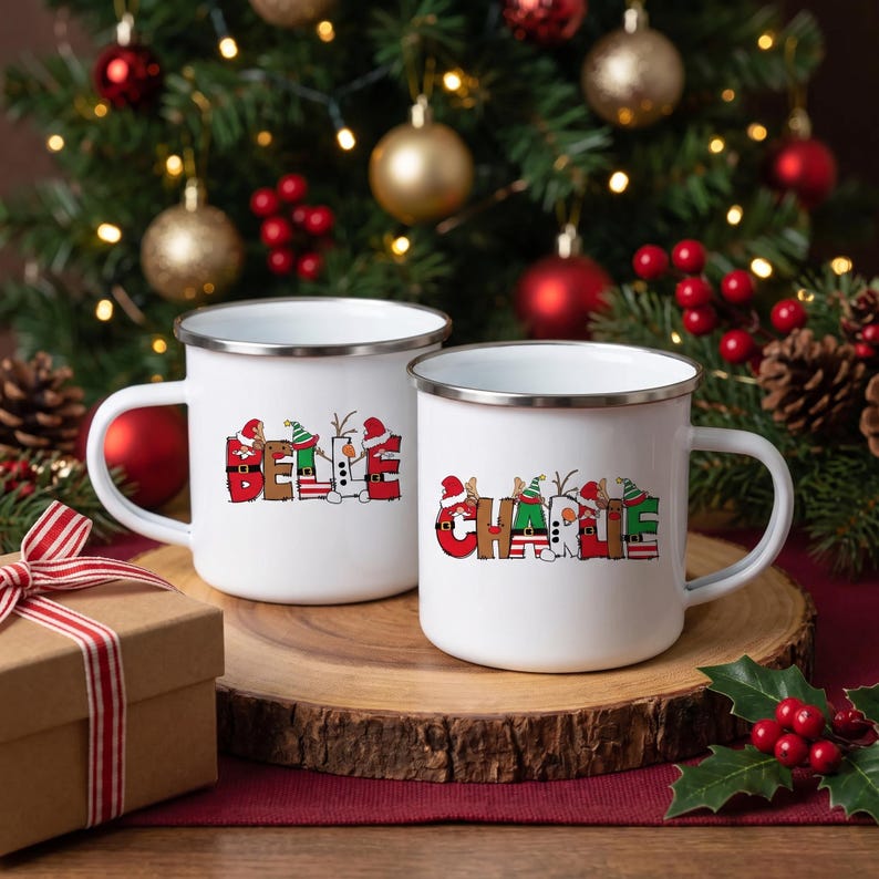 K&ouml;nnte beinhalten: Zwei wei&szlig;e Emaille-Tassen mit silbernen R&auml;ndern. Die linke Tasse zeigt den Namen BELLE in roten, gr&uuml;nen und wei&szlig;en Buchstaben im Feiertagsdesign. Die rechte Tasse zeigt den Namen CHARLIE in &auml;hnlicher festlicher Schrift mit Weihnachts- und Elfenmotiven.