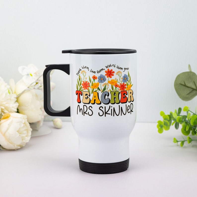 May include: White travel mug with a black handle, lid, and base. The mug features the text "TEACHER MRS SKINNER" in colorful letters, with a floral design above the text. The words "Teach them, love them, watch them grow" are printed above the flowers.
