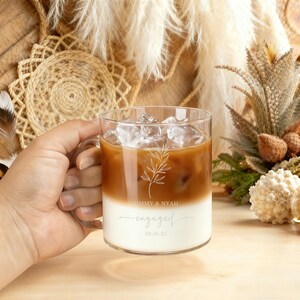 Peut inclure: Un mug en verre transparent rempli de caf&eacute; glac&eacute;, avec une base blanche cr&eacute;meuse et une couche sup&eacute;rieure marron. Le mug a une anse et est d&eacute;cor&eacute; d'un motif floral et des mots "engaged" et "10.10.25".
