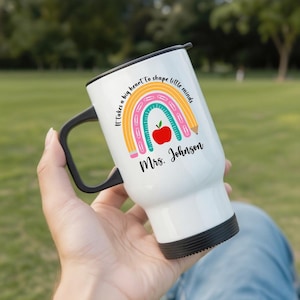 Puede incluir: Taza de viaje blanca con asa y tapa negras. La taza presenta un colorido dise&ntilde;o de arco&iacute;ris con una manzana y un l&aacute;piz, y el texto "It takes a big heart to shape little minds" y "Mrs. Johnson".