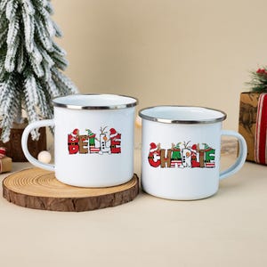 K&ouml;nnte beinhalten: Zwei wei&szlig;e Emaille-Tassen mit silbernen R&auml;ndern. Jede Tasse ist mit einem personalisierten Namen in einer festlichen Schriftart bedruckt: eine zeigt BELLE und die andere CHARLIE. Die Buchstaben sind mit weihnachtlichen Motiven wie Weihnachtsmannm&uuml;tzen, Rentiergeweihen und Streifenmustern verziert.
