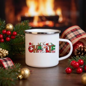 K&ouml;nnte beinhalten: Ein wei&szlig;er Emaillebecher mit silbernem Rand, der den Namen CHARLIE in bunten, weihnachtlichen Blockbuchstaben zeigt. Jeder Buchstabe ist mit festlichen Elementen wie Weihnachtsmannm&uuml;tzen, Rentiergeweihen und Streifenmustern verziert.