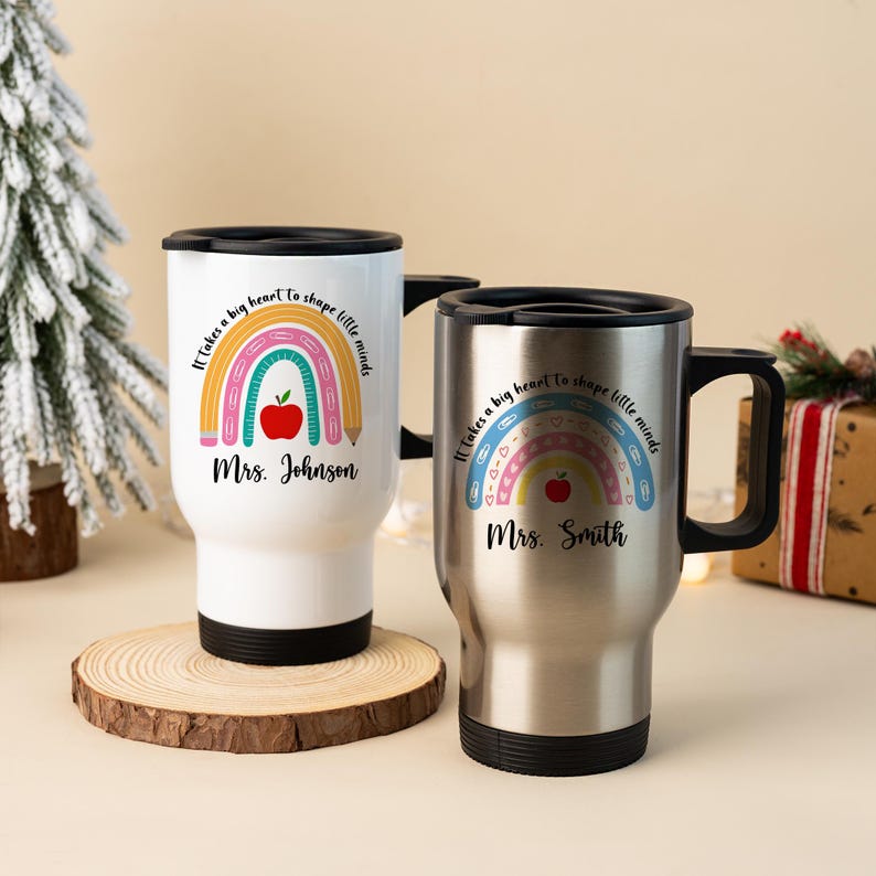 Puede incluir: Dos tazas de viaje, una blanca y otra plateada, con tapas y asas negras. Cada taza presenta un dise&ntilde;o de arco&iacute;ris con una manzana y un l&aacute;piz, y el texto "It takes a big heart to shape little minds" y un nombre.