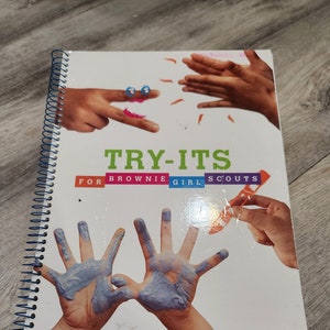Puede incluir: Un libro blanco con una encuadernación en espiral azul. La portada presenta el texto "TRY-ITS FOR BROWNIE GIRL SCOUTS" e imágenes de manos de niños con pintura.