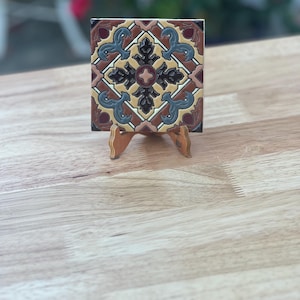May include: A square decorative tile with a detailed, symmetrical pattern in shades of brown, tan, blue, and burgundy. The tile is displayed on a small wooden easel, showcasing its design. The tile's dimensions are approximately 4 inches by 4 inches.