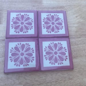 May include: Four square ceramic coasters with a floral design. Each coaster has a light purple background with a white square in the center. A light purple flower design is in the center of the white square.