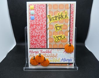 May include: A handmade Thanksgiving card with the words "Thankful for you" in yellow lettering. The card features a red floral pattern, pumpkin illustrations, and small orange pumpkin embellishments. The phrase "Always Thankful" is also visible.