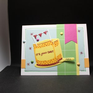 May include: A birthday card with a yellow cake with brown frosting and the text "It's your day!" on it. The cake is decorated with red, white, and blue flags. The card has a green and white checkered background.