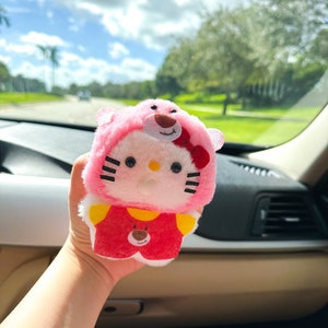 May include: Pink Hello Kitty plush toy wearing a pink bear costume with a red bow. The toy is being held in a person's hand.