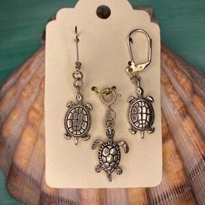 May include: A set of three silver turtle charms on a white card. Two of the charms are earrings and the third is a charm with a lobster clasp.