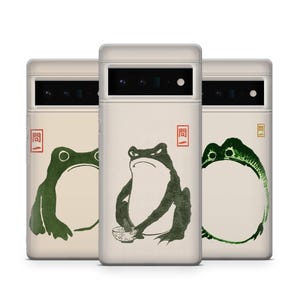 May include: Three phone cases with a cream background and a Japanese-style frog illustration in green. Each case features a different frog design, with one holding a bowl. The cases also have a red square with a Japanese character.