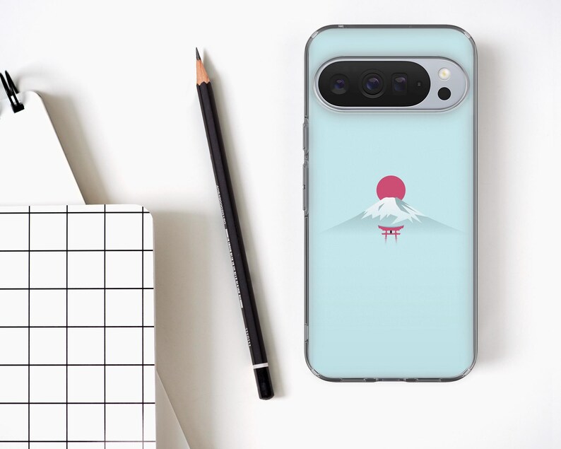 May include: A light blue phone case with a minimalist design of a mountain, a red sun, and a Japanese torii gate. A black pencil and a white notebook with a grid pattern are also visible.