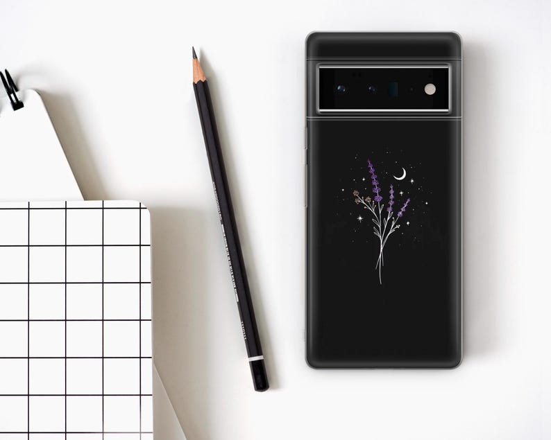 May include: Black phone case featuring a floral design with lavender and a crescent moon. The case is next to a black pencil and a white notebook with a grid pattern. The phone case has a camera and flash.