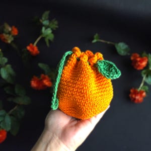 Puede incluir: Una pequeña bolsa de cordón de ganchillo naranja con una hoja de ganchillo verde adjunta. La bolsa se sostiene en una mano.