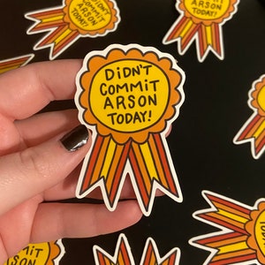 May include: A yellow and orange ribbon sticker with the text "Didn't Commit Arson Today!" in black lettering. The sticker is shaped like a ribbon with a circular centre.