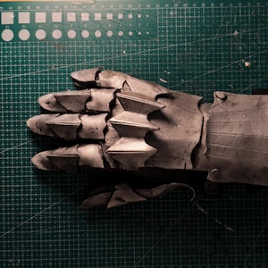 May include: A detailed, metallic-looking gauntlet, likely for cosplay or costume use. The gauntlet is a muted silver color, with articulated fingers and a segmented design. The surface appears textured, suggesting a weathered or aged appearance.