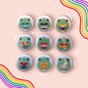 Puede incluir: Nueve botones redondos blancos con ilustraciones de ranas verdes que sostienen corazones con diferentes banderas de orgullo. Las banderas incluyen las banderas bisexual, transgénero, lesbiana, pansexual, genderqueer, no binario y asexual.