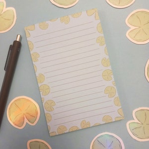 Puede incluir: Un bloc de notas azul con líneas y un borde de nenúfares verdes. El bloc de notas está sobre un fondo azul claro con varias pegatinas de nenúfares verdes.