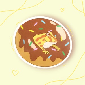 May include: A yellow and brown frog sits on top of a chocolate doughnut with colourful sprinkles. The doughnut is on a white background.