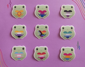 May include: Nine frog stickers with different pride flags on their hearts. The flags include the bisexual, lesbian, transgender, non-binary, pansexual, genderfluid, and asexual flags.