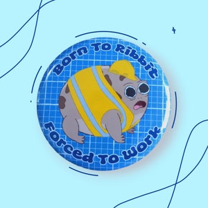 May include: A blue and white button with a cartoon frog wearing a yellow construction vest and a hard hat. The text on the button reads "Born to Ribbit Forced to Work".