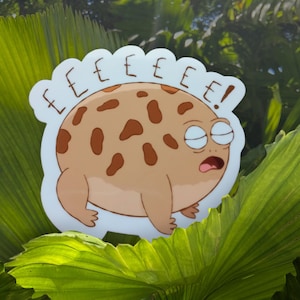 May include: A sticker featuring a cartoon frog with a light brown body and dark brown spots. The frog has closed eyes and an open mouth, with the word "EEEEEE!" above its head. The sticker is set against a backdrop of green leaves.