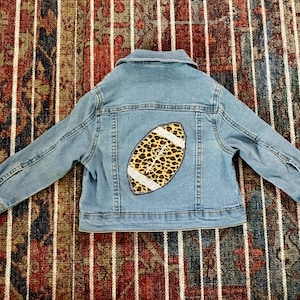 May include: A light blue denim jacket featuring a long-sleeved design with a collar and buttoned cuffs. The back panel displays a large, centered applique of an American football with a brown and black leopard print and white sequin details.