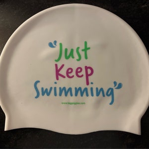 May include: White silicone swim cap with the text "Just Keep Swimming" in green, pink, and blue lettering. The text is enclosed in blue quotation marks. The website "www.taggoggles.com" is printed in black below the text.