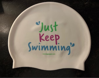 May include: White silicone swim cap with the text "Just Keep Swimming" in green, pink, and blue lettering. The text is enclosed in blue quotation marks. The website "www.taggoggles.com" is printed in black below the text.
