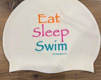 May include: White silicone swim cap with the text "Eat Sleep Swim" in orange, pink, and teal lettering. The text "www.tagoggles.com" is printed in black below the text.