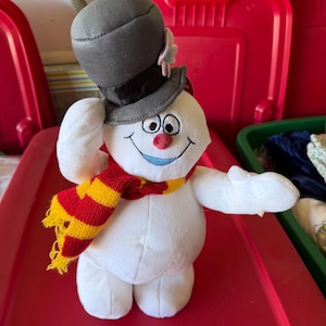 May include: A plush snowman toy with a black top hat, red and yellow scarf, and a red nose. The snowman has a smiling face and is set against a red background. The snowman is a winter holiday decoration.