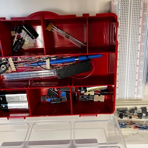 May include: A red plastic organizer box filled with electronic components including resistors, capacitors, integrated circuits, a battery holder, and two AA batteries. Beside the box are two white breadboards and a clear bag of assorted wires.