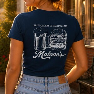 May include: A navy blue short-sleeve t-shirt featuring a white graphic on the back. The design includes a beer mug, a burger, and the text "BEST BURGERS IN HASTINGS, MA" above the word "Malone's" in a script font. The shirt is paired with light blue denim shorts.
