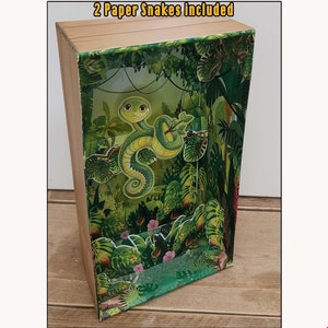 May include: A papercraft diorama of a jungle terrarium with a green snake inside. The diorama is made of cardboard and has a wooden frame. The text "Snake Tank - Jungle Terrarium - Paper Diorama" and "2 Paper Snakes Included" is printed on the top of the diorama.