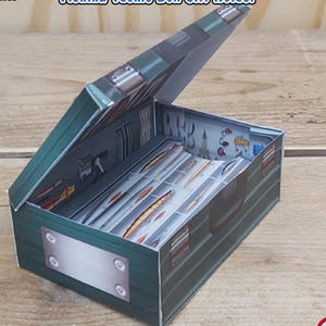 Peut inclure: Une boîte cadeau en papier vert en forme de boîte à pêche avec le texte "Fishing Tackle Box Gift Holder". La boîte est ouverte, révélant un compartiment avec des leurres de pêche. Le texte "Digital Download - DIY Paper Tackle Box with Gift Compartment" est également visible.