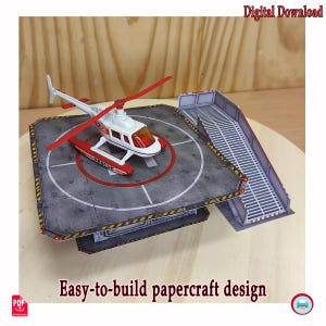 May include: A papercraft model of a white and red rescue helicopter sitting on a gray helipad platform with a matching staircase. The text Digital Download and Easy-to-build papercraft design are visible along with a PDF icon and a printer icon.