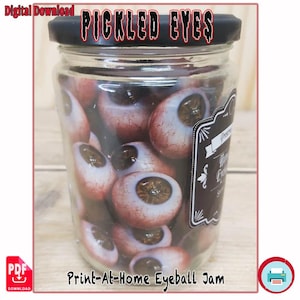 Peut inclure: Un bocal en verre rempli d'yeux conservés. Le bocal a un couvercle noir et une étiquette. Le texte "PICKLED EYES" est en haut et "Print-At-Home Eyeball Jam" en bas. Les globes oculaires sont bruns et blancs.