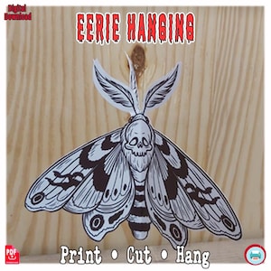 Könnte beinhalten: Schwarz-weiße Illustration einer Motte mit Totenkopfgesicht und Fledermausflügelmustern. Der Text "EERIE HANGING" steht oben und "Print • Cut • Hang" unten. Das Bild befindet sich auf einem hellen Holzgrund.