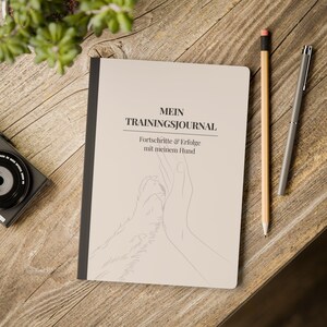 Può includere: Un diario di allenamento beige con il testo "MEIN TRAININGSJOURNAL" e "Fortschritte & Erfolge mit meinem Hund" è aperto su una superficie di legno. Una matita e una penna sono accanto al diario. Una fotocamera e una pianta sono sullo sfondo.