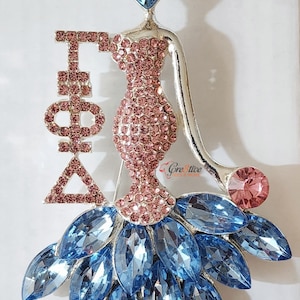 May include: A silver pin with a pink and blue crystal design. The pin features a woman in a dress with a large crystal headpiece. The letters "Iota Phi Delta" are on the left side of the pin.