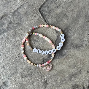 Puede incluir: Dos pulseras de cuentas con cuentas rosas, blancas y grises. Una pulsera tiene el nombre "ISAIAH" escrito en letras blancas sobre cuentas negras. La otra pulsera tiene el número "410" escrito en letras blancas sobre cuentas negras. Ambas pulseras tienen un pequeño dije de mariposa rosa.