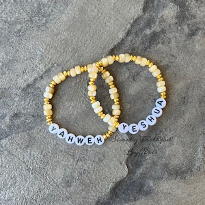 May include: Two beaded bracelets with the words "YAHWEH" and "YESHUA" spelled out in black letters on white beads. The bracelets have gold-colored accents and are made with cream-colored beads. The bracelets are displayed on a gray surface.