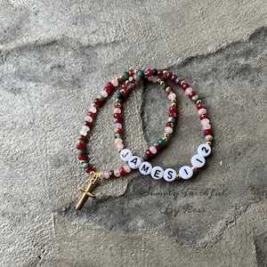 May include: Two beaded bracelets with a gold cross charm. One bracelet spells out "JAMES" and the other spells out "112".