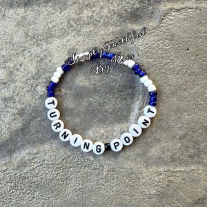 May include: A bracelet made of white letter beads spelling "TURNING POINT", interspersed with blue and white beads. The text "Simply Faithful By Rois" is written in cursive above the bracelet.
