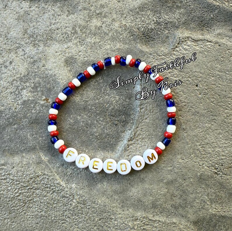 May include: A bracelet made of red, white, and blue beads. The word "FREEDOM" is spelled out in white letter beads with gold lettering. The bracelet is displayed on a grey surface. The text "Simply Faithful By Kris" is in the background.