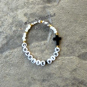 May include: A beaded bracelet spelling out "CHARLIE KIRK" in white letter beads. The bracelet has white and gold beads, a black cross, and the text "Simply Faithful By Kris".