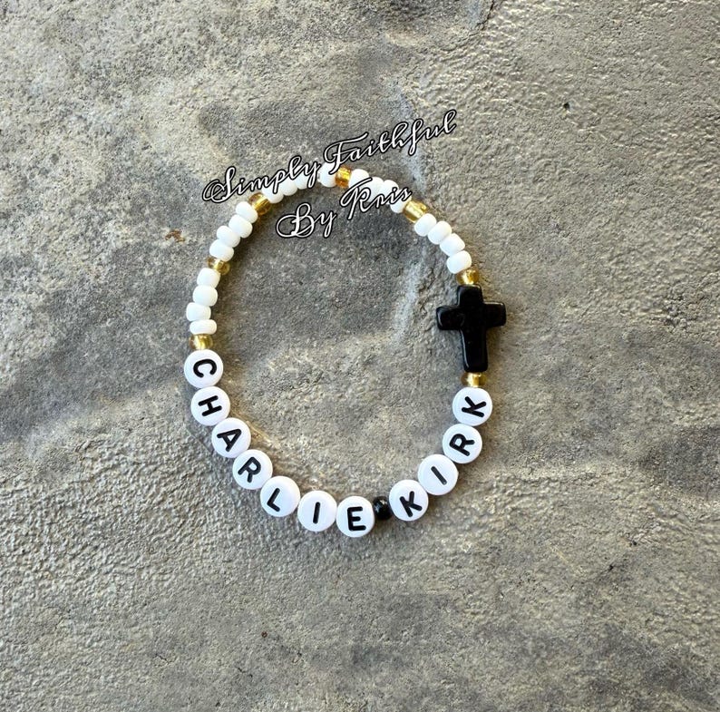 May include: A beaded bracelet spelling out "CHARLIE KIRK" in white letter beads. The bracelet has white and gold beads, a black cross, and the text "Simply Faithful By Kris".